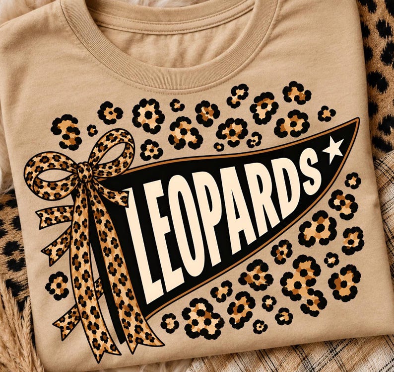 May include: A tan t-shirt featuring a black pennant graphic with the word LEOPARDS in white capital letters. The design includes a leopard print bow on the left side of the pennant, a white star on the right, and leopard print spots scattered around.
