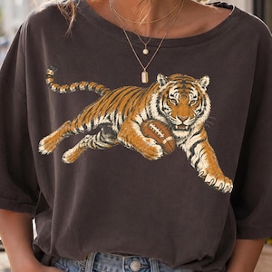 May include: A charcoal gray short-sleeve t-shirt featuring a graphic print of a tiger in mid-stride, holding a brown American football. The tiger is orange with black stripes and white accents. The shirt has a wide crew neckline and loose fit.