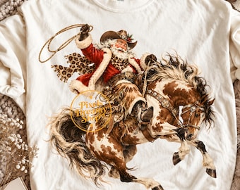 May include: An off-white crewneck t-shirt featuring a graphic of Santa Claus as a cowboy riding a brown and white spotted horse. Santa wears a red coat, a cowboy hat, and a leopard print scarf while holding a lasso. The text Pixels Dreamy Shop is visible.