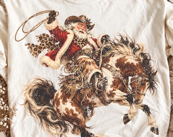 May include: An off-white crewneck t-shirt featuring a graphic of Santa Claus wearing a cowboy hat and a leopard print scarf while riding a brown and white spotted horse. Santa is holding a lasso in the air. The shirt has a Comfort Colors label.