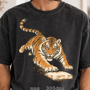May include: A charcoal black, acid-washed t-shirt featuring a graphic illustration of a tiger in a pouncing position with its front paws resting on a rectangular block. The tiger is orange with black stripes and white accents on its face.
