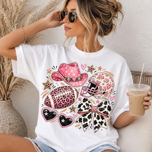 May include: A white short-sleeve t-shirt featuring a graphic print of a pink cowboy hat, a leopard print football, a floral football helmet, heart-shaped sunglasses, and a cow-print bow, all accented with small stars and hearts.
