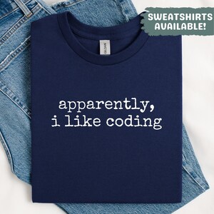 Puede incluir: Camiseta azul marino con el texto blanco "apparently, i like coding". La camiseta está doblada y colocada sobre unos vaqueros azules. Un recuadro verde en la esquina superior derecha dice "SWEATSHIRTS AVAILABLE!"