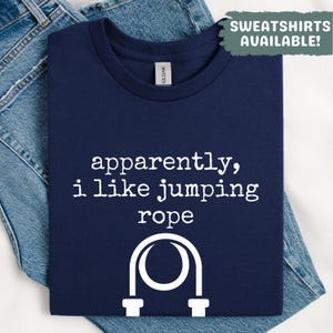 Op de afbeelding: Marineblauw T-shirt met de witte tekst "apparently, i like jumping rope" en een afbeelding van een springtouw. Het T-shirt is op een blauwe jeans gevouwen. De tekst "SWEATSHIRTS AVAILABLE!" staat rechtsboven.
