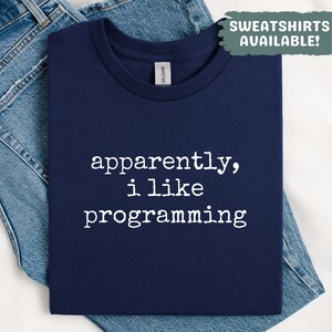 May include: Navy blue t-shirt with the text "apparently, i like programming" in white. The shirt is folded and placed on a pair of blue jeans. The top right corner has a green box that says "SWEATSHIRTS AVAILABLE!"