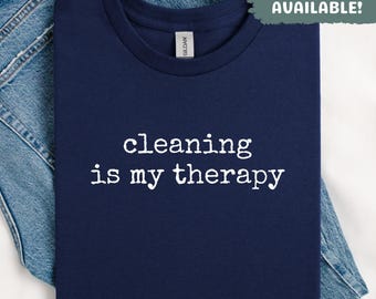 May include: Navy blue t-shirt with the white text "cleaning is my therapy." The shirt is folded on top of a pair of blue jeans. The text "SWEATSHIRTS AVAILABLE!" is in a green box in the upper right corner.