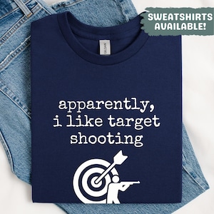 May include: Navy blue t-shirt with the text "apparently, i like target shooting" in white. A graphic of a target with an arrow and a person shooting a gun is below the text. The shirt is folded on top of blue denim jeans.