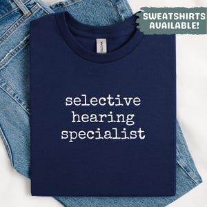 May include: A navy blue t-shirt with the text "selective hearing specialist" in white. The shirt is folded and placed on top of a pair of blue jeans. The text "SWEATSHIRTS AVAILABLE!" is in a green box.
