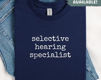 Puede incluir: Una camiseta azul marino con el texto "selective hearing specialist" en blanco. La camiseta está doblada y colocada encima de unos vaqueros azules. El texto "SWEATSHIRTS AVAILABLE!" está en un cuadro verde.