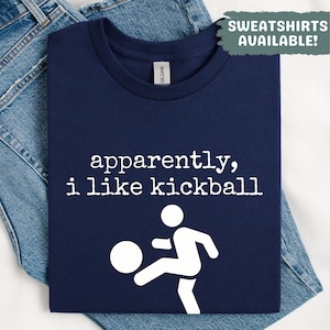 Op de afbeelding: Marineblauw T-shirt met de witte tekst "apparently, i like kickball". Een witte afbeelding van een persoon die tegen een bal trapt staat eronder. Het T-shirt ligt op een blauwe spijkerbroek.
