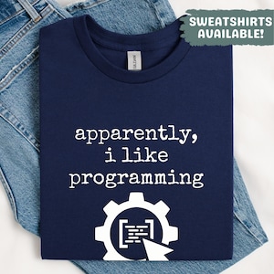 Può includere: T-shirt blu navy con la scritta bianca "apparently, i like programming". Sotto c'è una grafica bianca di un ingranaggio con codice e un cursore. La maglietta è piegata sopra dei jeans blu. Il testo "SWEATSHIRTS AVAILABLE!" è in alto a destra.