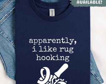 Puede incluir: Camiseta azul marino con el texto blanco "apparently, i like rug hooking" y un gráfico de herramientas para hacer ganchillo de alfombras. La camiseta está doblada sobre unos vaqueros azules. Arriba a la derecha pone "Sweatshirts Available!"