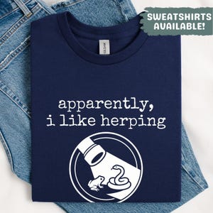 Może przedstawiać: Granatowa koszulka z napisem "apparently, i like herping" i białą grafiką węża i żaby. Koszulka jest złożona na niebieskich dżinsach. Słowa "SWEATSHIRTS AVAILABLE!" znajdują się w prawym górnym rogu.