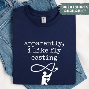 Könnte beinhalten: Marineblaues T-Shirt mit dem weißen Text "apparently, i like fly casting". Darunter befindet sich eine weiße Grafik einer angelnden Person. Das T-Shirt liegt auf einer blauen Jeans. Der Text "SWEATSHIRTS AVAILABLE!" ist oben rechts.