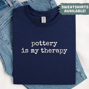 Może przedstawiać: Granatowa koszulka z białym napisem "pottery is my therapy". Koszulka jest złożona na niebieskich dżinsach. Zielono-białe pole tekstowe w prawym górnym rogu mówi "SWEATSHIRTS AVAILABLE!"