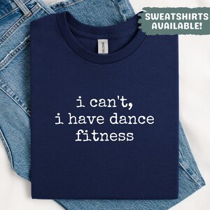May include: Navy blue t-shirt with the text "i can't, i have dance fitness" in white. The shirt is folded on top of a pair of blue jeans. The words "SWEATSHIRTS AVAILABLE!" are in a green box in the upper right corner.