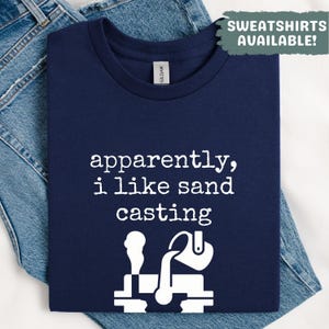 May include: Navy blue t-shirt with the text "apparently, i like sand casting" in white, along with a graphic of a sand casting process. The shirt is folded on top of blue denim jeans. The text "SWEATSHIRTS AVAILABLE!" is in the upper right corner.