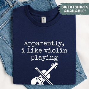 May include: A navy blue t-shirt featuring the white text "apparently, i like violin playing" and a white violin graphic. The shirt is folded on denim jeans. A green box says "SWEATSHIRTS AVAILABLE!"