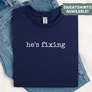 May include: Navy blue t-shirt with the phrase "he's fixing" in white, centered on the front. The shirt is folded on top of blue denim jeans. A green banner in the upper right corner reads "SWEATSHIRTS AVAILABLE!"