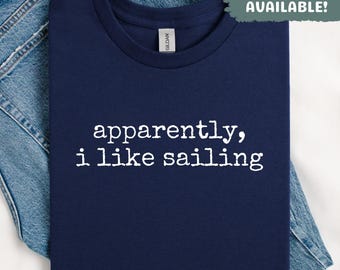 May include: Navy blue t-shirt with the text "apparently, i like sailing" in white. The shirt is folded and placed on top of a pair of blue jeans. The words "SWEATSHIRTS AVAILABLE!" are in a green box.