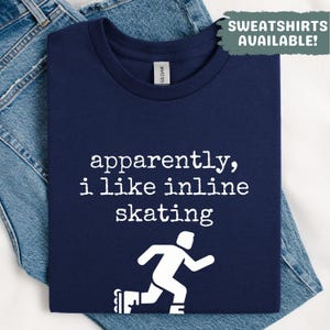 May include: Navy blue t-shirt with the text "apparently, i like inline skating" in white. A white graphic of a person inline skating is below the text. The shirt is folded on top of blue jeans. The text "SWEATSHIRTS AVAILABLE!" is in the upper right corner.