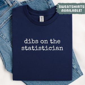 Puede incluir: Camiseta azul marino con la frase blanca "dibs on the statistician". Doblada sobre unos vaqueros azules. En la esquina superior derecha, un recuadro verde dice "SWEATSHIRTS AVAILABLE!".