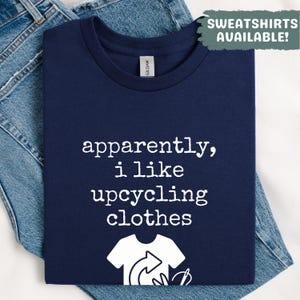May include: Navy blue t-shirt with the text "apparently, i like upcycling clothes" in white. The shirt features a graphic of a t-shirt with a recycling symbol. The words "Sweatshirts Available!" are in a green box.