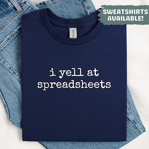 May include: A navy blue t-shirt with the white text "i yell at spreadsheets". The t-shirt is folded on a pair of blue jeans. A green box in the top right corner reads "SWEATSHIRTS AVAILABLE!"