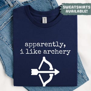 May include: Navy blue t-shirt with the text "apparently, i like archery" and a white bow and arrow graphic. The shirt is folded on top of blue denim jeans. The words "SWEATSHIRTS AVAILABLE!" are in a green box in the upper right corner.