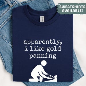 May include: Navy blue t-shirt with the text "apparently, i like gold panning" in white. A white graphic of a person panning for gold is below the text. The shirt is folded on top of blue denim jeans.