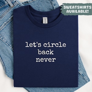 May include: Navy blue t-shirt with the text "let's circle back never" in white. The shirt is folded on top of a pair of blue jeans. The words "SWEATSHIRTS AVAILABLE!" are in a green box in the upper right corner.