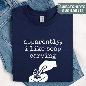 Pode incluir: Camiseta azul marinho com o texto branco "apparently, i like soap carving". Um gráfico branco mostra uma mão esculpindo uma flor em sabão. A camiseta está dobrada sobre um par de jeans azuis.