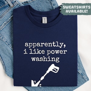 Può includere: T-shirt blu navy con la scritta "apparently, i like power washing" e un disegno bianco di un'idropulitrice. La maglietta è piegata sopra dei jeans blu. Una scatola verde dice "Sweatshirts Available!"