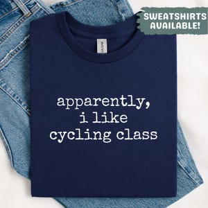 May include: Navy blue t-shirt with the text "apparently, i like cycling class" in white. The shirt is folded on top of a pair of blue jeans. The top right corner of the image has a green box with the text "SWEATSHIRTS AVAILABLE!"