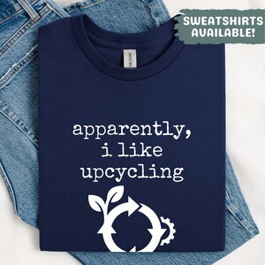May include: A navy blue t-shirt with the white text "apparently, i like upcycling". It has a white recycling symbol graphic with leaves and a gear. A green box says "SWEATSHIRTS AVAILABLE!"