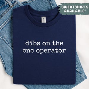 Op de afbeelding: Marineblauw T-shirt met de witte tekst "dibs on the cnc operator". Het T-shirt is opgevouwen op een blauwe jeans. Een groene box in de rechterbovenhoek toont "SWEATSHIRTS AVAILABLE!"