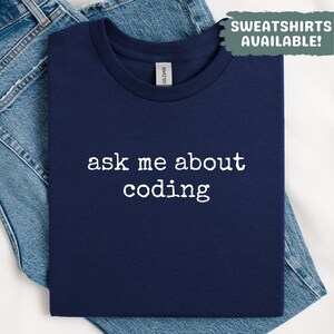 May include: A navy blue t-shirt with the text "ask me about coding" in white. The shirt is folded and placed on top of a pair of blue jeans. The image also includes a green box that says "SWEATSHIRTS AVAILABLE!"