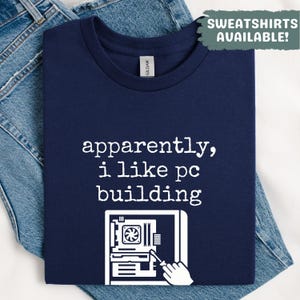 Könnte beinhalten: Marineblaues T-Shirt mit dem weißen Text "apparently, i like pc building". Die Grafik zeigt eine Computerkomponente und eine Hand mit einem Schraubendreher. Das T-Shirt liegt auf einer blauen Jeans.
