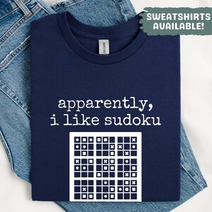Può includere: T-shirt blu navy con la scritta "apparently, i like sudoku" e un motivo a puzzle Sudoku bianco. La maglietta è piegata su una superficie di jeans. Il testo "SWEATSHIRTS AVAILABLE!" è in alto a destra. Realizzato in materiale morbido.