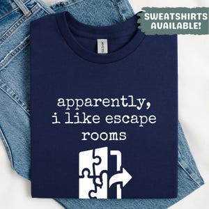 May include: Navy blue t-shirt with the text "apparently, i like escape rooms" in white. A white graphic of puzzle pieces and an arrow is below the text. The shirt is folded on top of blue denim jeans. The text "SWEATSHIRTS AVAILABLE!" is in the top right corner.