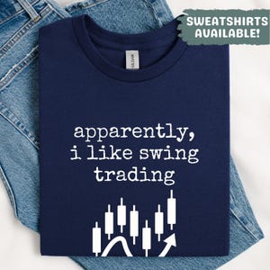May include: Navy blue t-shirt with the text "apparently, i like swing trading" in white. Below the text is a graphic of stock market candlesticks and an upward arrow. The shirt is folded on top of blue jeans.