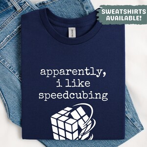May include: Navy blue t-shirt with the text "apparently, i like speedcubing" in white. A white graphic of a Rubik's Cube is below the text. The shirt is folded on top of blue denim jeans.