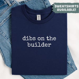 May include: A navy blue t-shirt with the phrase "dibs on the builder" printed in white. The shirt is folded on top of a pair of blue jeans. The text "SWEATSHIRTS AVAILABLE!" is in the upper right corner.