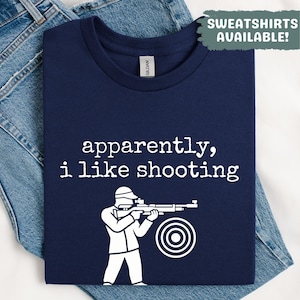 May include: Navy blue t-shirt with the text "apparently, i like shooting" and a white graphic of a person aiming a rifle at a target. The shirt also has the text "SWEATSHIRTS AVAILABLE!"