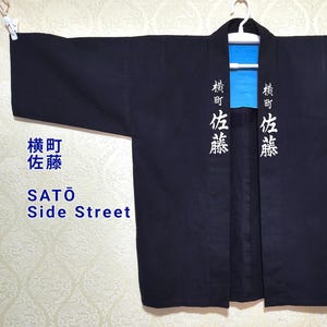 Puede incluir: Una chaqueta japonesa azul marino oscuro estilo kimono con caracteres japoneses blancos y el texto en inglés "SATŌ Side Street" impreso en la parte delantera. El forro interior es de color azul brillante. La chaqueta está colgada en una percha blanca.