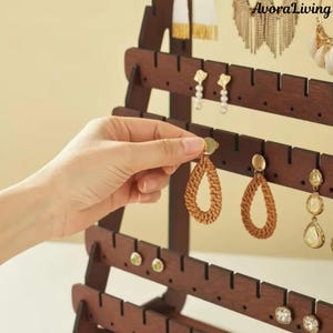 May include: A dark brown wooden earring display stand with multiple rows of slots for hanging earrings. A hand is placing a woven, teardrop-shaped earring on the stand. Other earrings of various styles are already displayed.