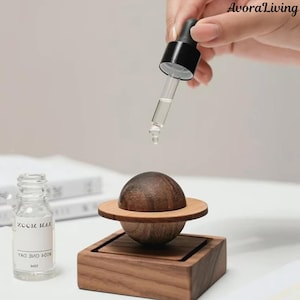 May include: A wooden essential oil diffuser with a planet design. The diffuser features a dark brown sphere with a light brown ring, set on a square wooden base. A glass bottle and dropper are in the image. The text "AvoraLiving" is in the upper right corner.