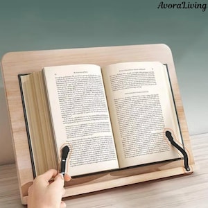 May include: A light wood book stand holding an open book. The stand has black page holders. The book is open to a page of text. The text "AvoraLiving" is visible in the top right corner. The stand is designed to hold books.