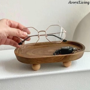 May include: A wooden tray with a natural finish, supported by three small wooden ball feet. A pair of gold-rimmed, octagonal eyeglasses rests on the tray, along with a black key fob. The brand name "AvoraLiving" is visible in the upper right corner.