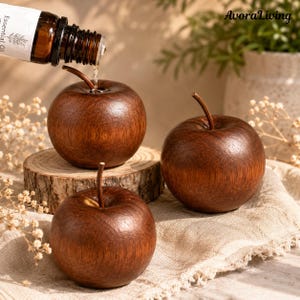 May include: Three wooden apple-shaped ornaments. One apple has essential oil being poured onto it from a brown glass bottle. The apples are a dark brown colour with a stem. The apples are on a wooden slice and a beige cloth.
